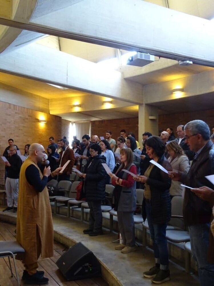 Grupo de personas de pie leyendo hojas en un auditorio, guiadas por un hombre con túnica amarilla y micrófono.