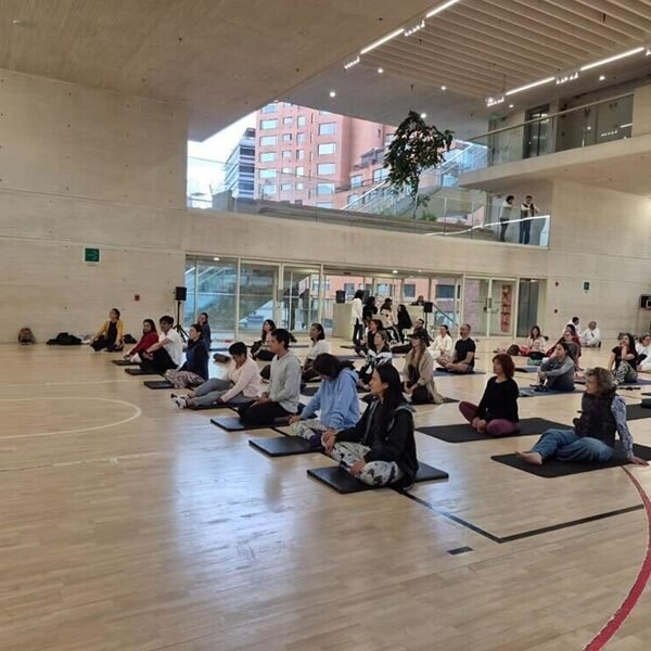 Grupo numeroso meditando sentado en el suelo de un gimnasio, guiado por un monje con túnica naranja.