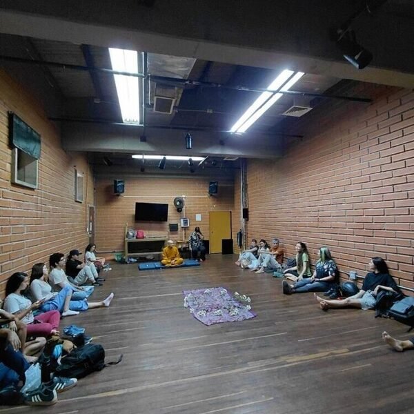 Grupo sentado en forma de U sobre un piso de madera, frente a un monje con túnica naranja en un salón de paredes de ladrillo.