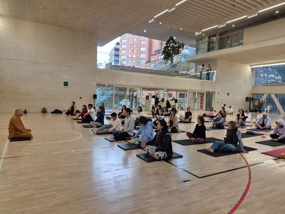Grupo numeroso meditando sentado en el suelo de un gimnasio, guiado por un monje con túnica naranja.