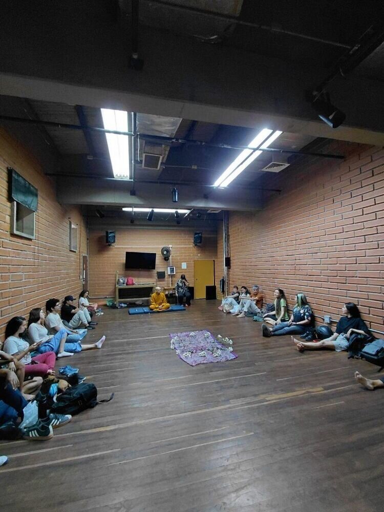 Grupo sentado en forma de U sobre un piso de madera, frente a un monje con túnica naranja en un salón de paredes de ladrillo.