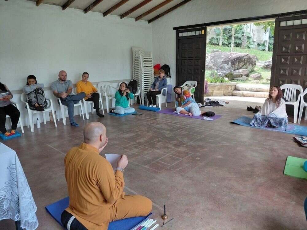 Sesión grupal en un salón con puertas abiertas al jardín, guiada por un monje sentado en una estera azul.