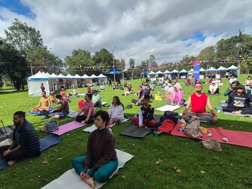 Decenas de personas meditando al aire libre sobre esterillas en un parque, bajo carpas blancas y banderines.