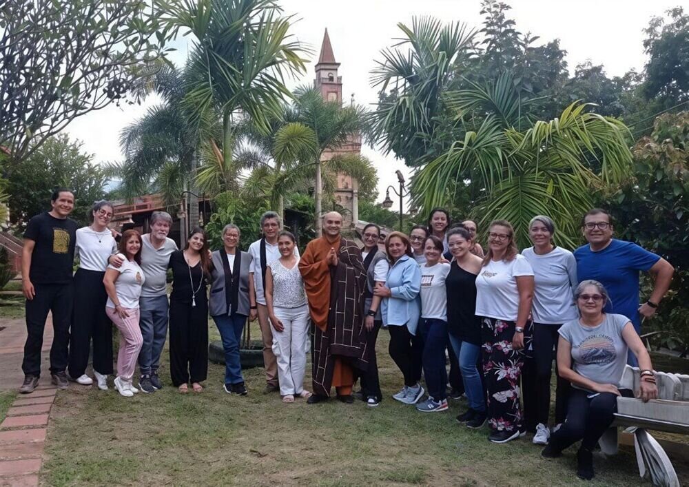 Grupo numeroso posando en un jardín junto a un monje con túnica marrón, con una torre de ladrillo al fondo.