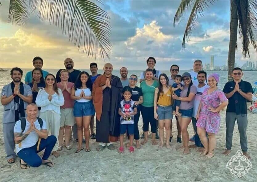 Grupo de la comunidad reunido en una playa al atardecer, con las manos juntas en gesto de saludo.