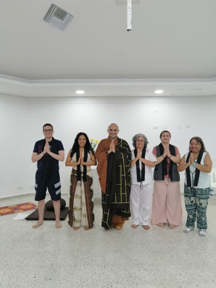 Seis personas de pie en una sala, con las manos juntas en gesto de meditación, junto a un monje.