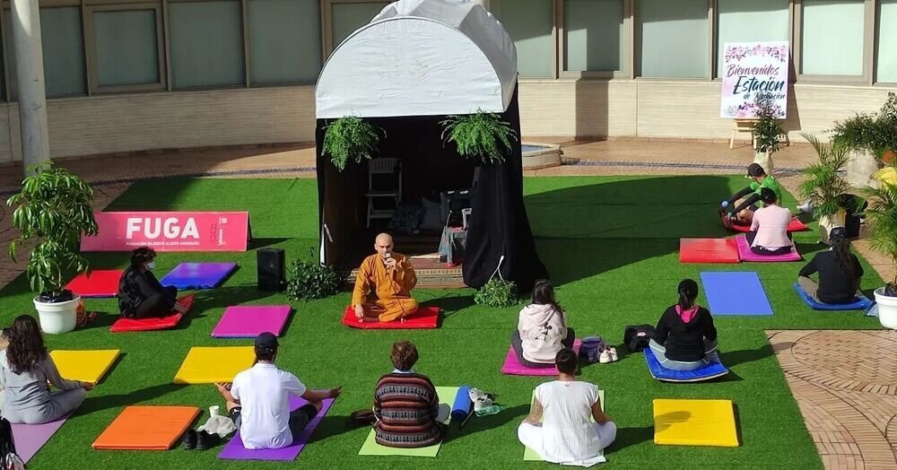 Grupo de personas meditando sentadas en cojines de colores sobre césped artificial, con un monje guiando la sesión bajo una carpa.