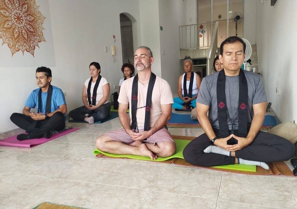 Grupo de personas meditando sentadas con los ojos cerrados en una sala, con un mural de mandala al fondo.
