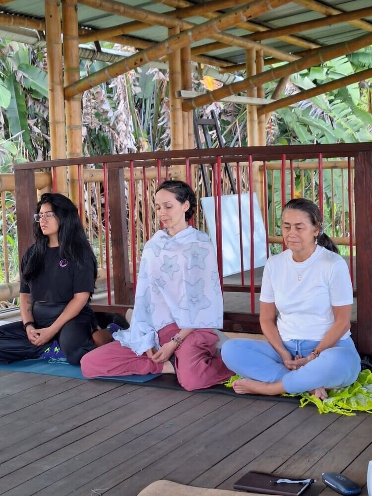 Tres mujeres meditando sentadas en el suelo bajo un techo de bambú, rodeadas de vegetación.