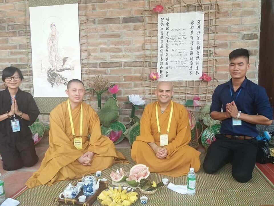 Dos monjes budistas sentados ante una mesa con té y frutas, acompañados de otras personas con las manos juntas, frente a un mural con caligrafía china.