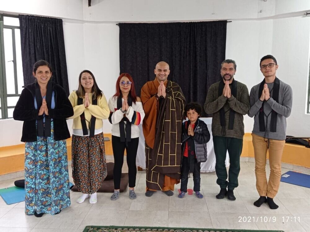 Grupo de siete personas y un niño con las manos juntas, junto a un monje, en una sala con cortinas oscuras.