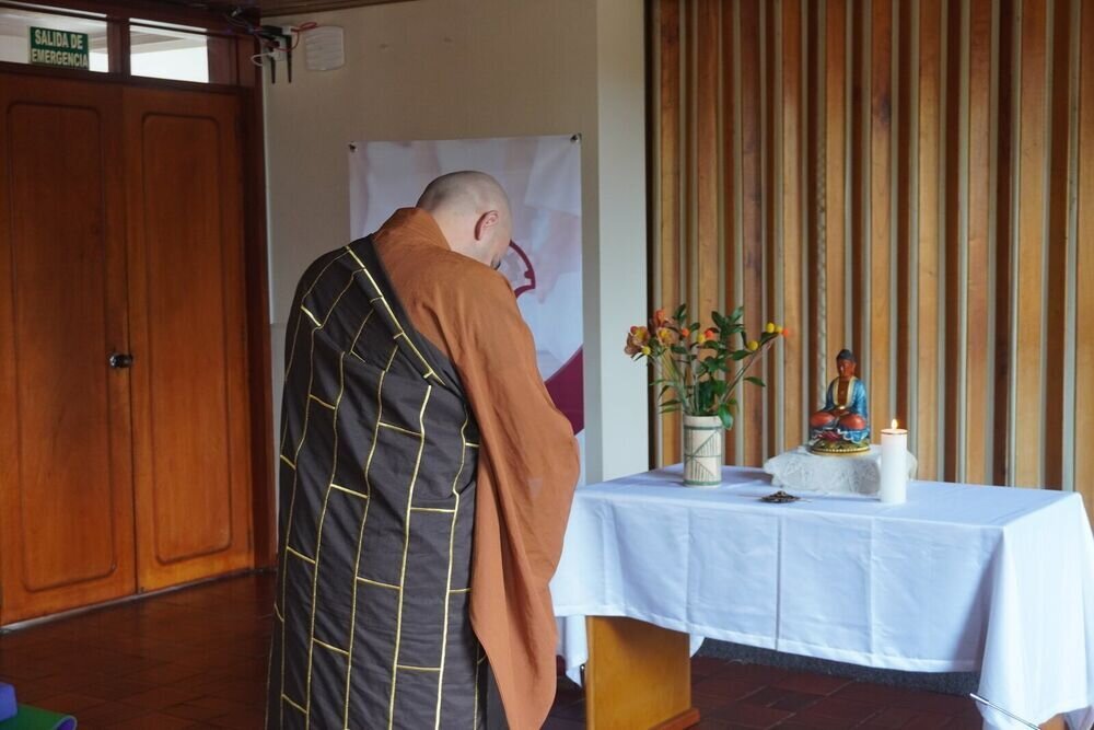 Un monje inclinado en reverencia ante un pequeño altar con una figura de Buda, flores y una vela encendida.