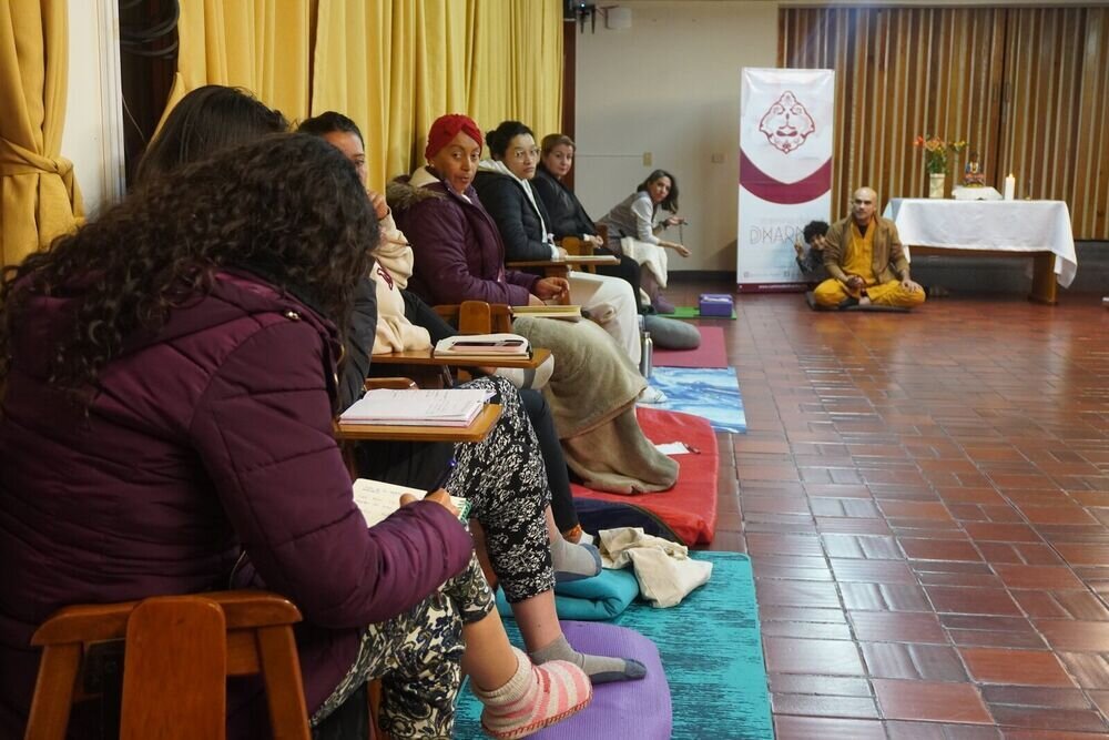 Grupo de personas sentadas en sillas y cojines en un salón, con un monje meditando al fondo junto a un altar con flores y una vela.