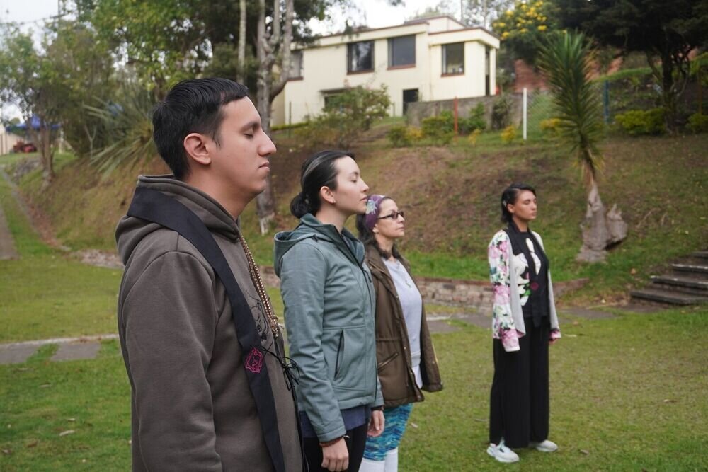 Cuatro personas de pie con los ojos cerrados en meditación, en un jardín frente a una casa.