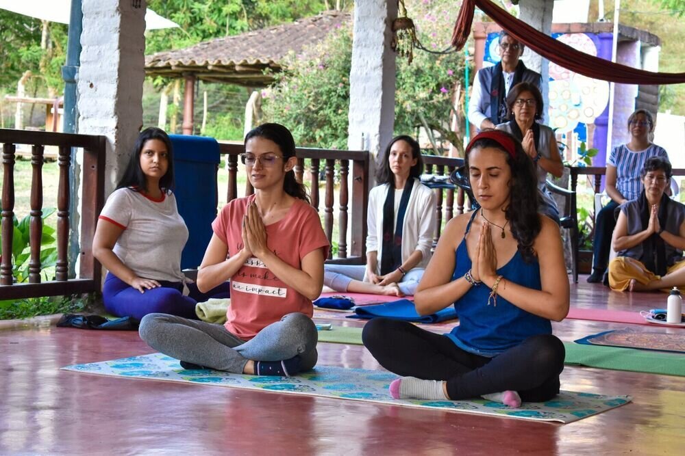 Grupo de personas meditando sentadas con las manos juntas, bajo un techo de tejas al aire libre.