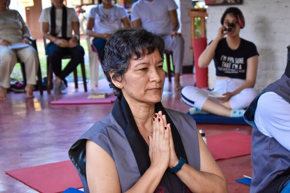 Una mujer meditando con las manos juntas en el pecho, rodeada de otras personas sentadas.