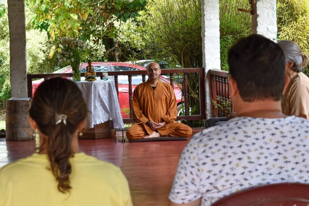Un monje meditando sentado frente a un altar con flores y una figura de Buda, con personas sentadas de espaldas en primer plano.